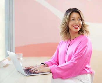 Smiling blonde woman in pink blouse working on laptop at desk in modern office